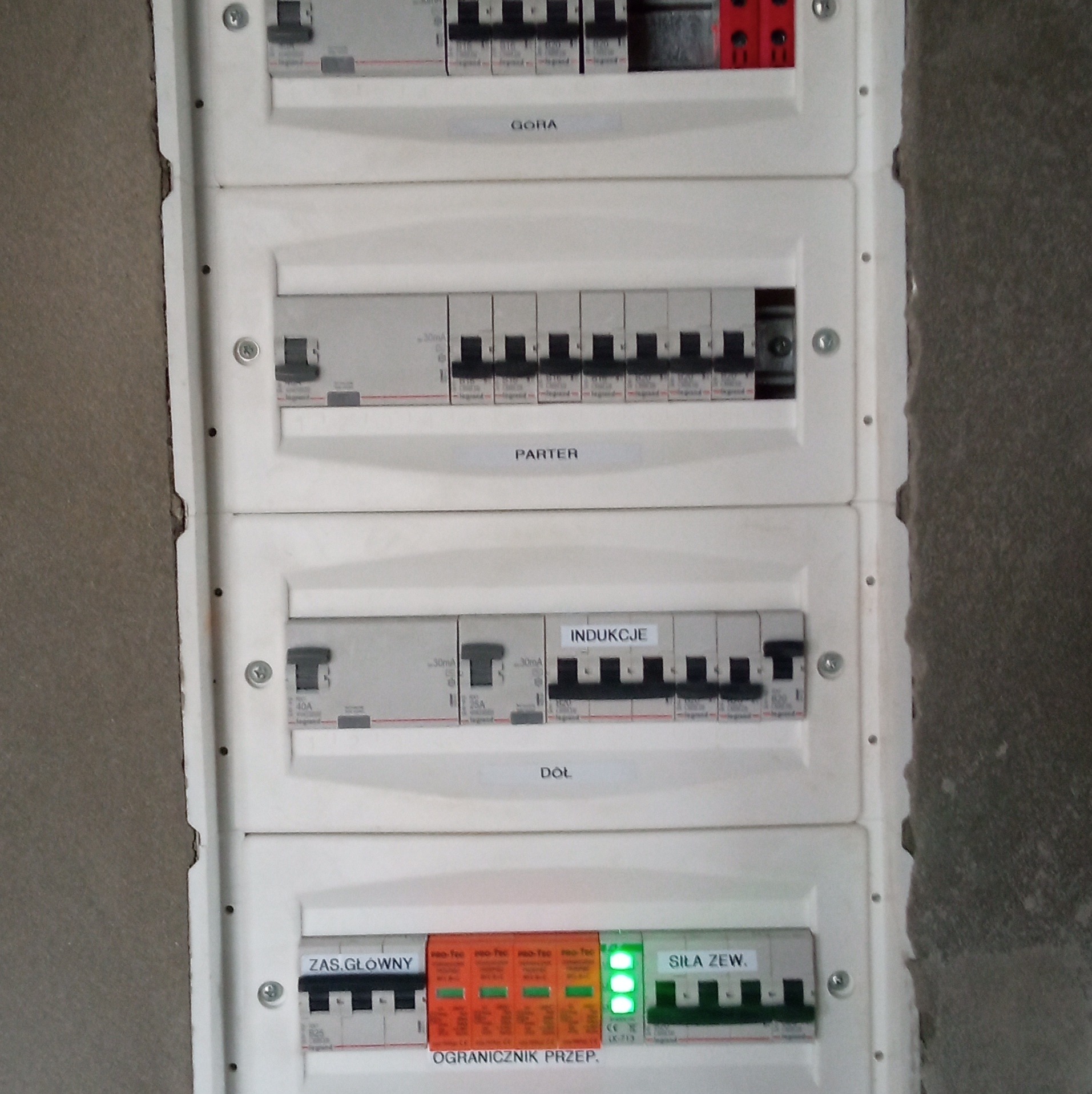 Rozdzielnica elektryczna z wyłącznikami różnicowoprądowymi i bezpiecznikami, opisana etykietami 'Góra', 'Parter', 'Indukcje', 'Dół', 'Zas. Główny', 'Siła Zew.', zamontowana na surowej ścianie.