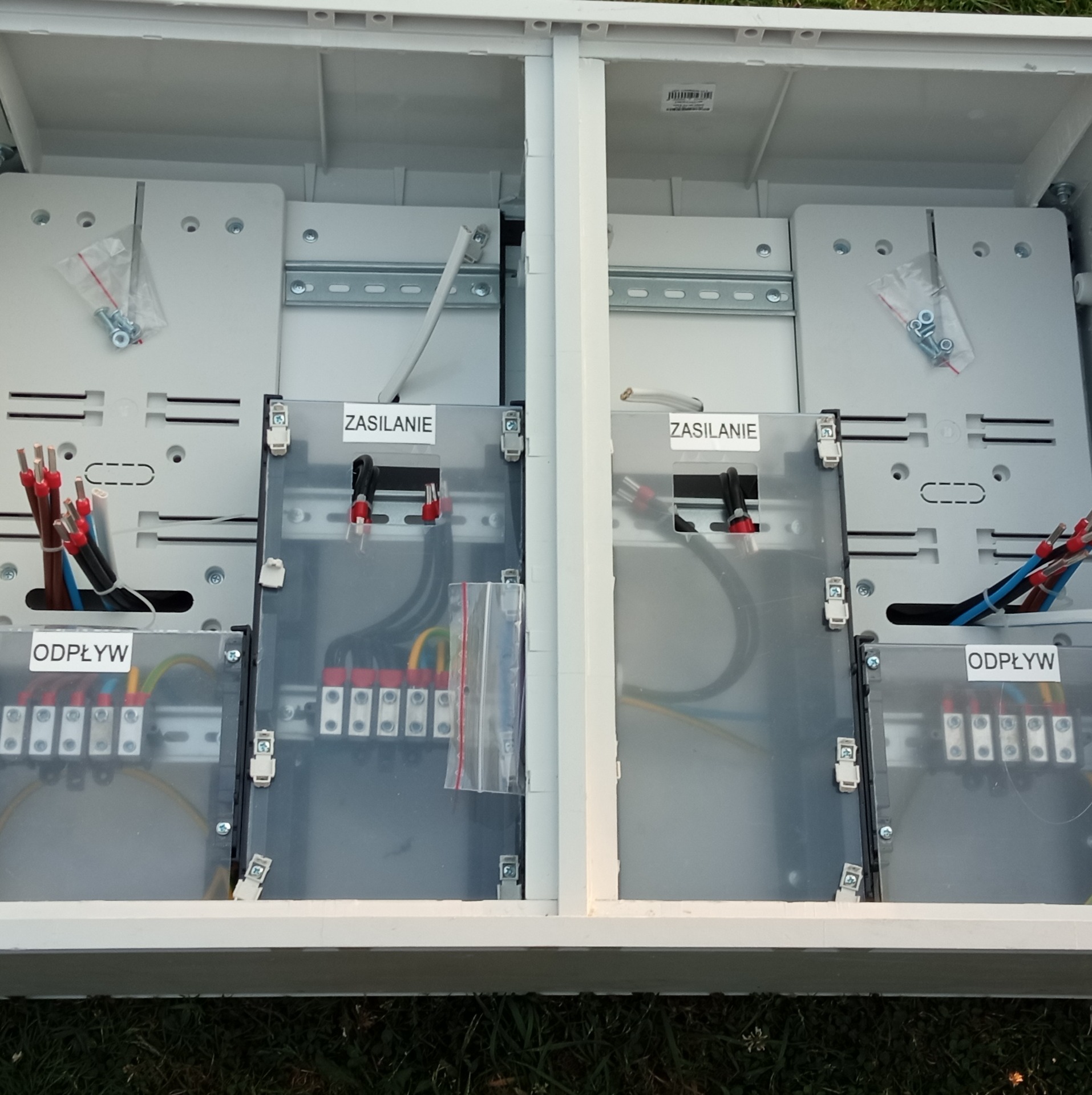 Otwarta, biała skrzynka elektryczna z widocznymi przewodami, zaciskami i etykietami 'Zasilanie' i 'Odpływ', umieszczona na trawie.