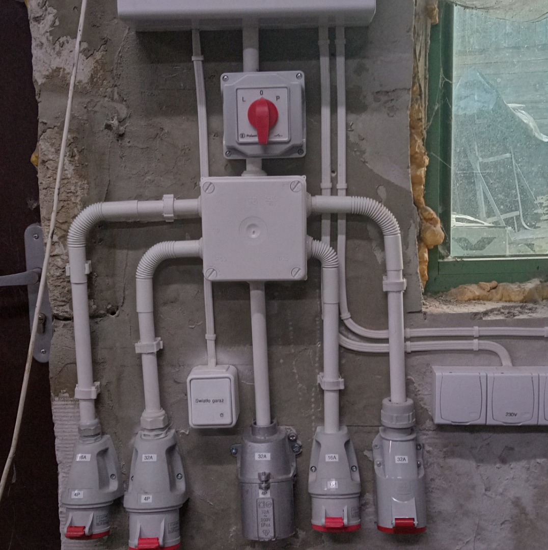 Rozdzielnica elektryczna z wyłącznikiem, zabezpieczeniami i gniazdami siłowymi na surowej ścianie, widoczne kable w peszlach i puszka 'światło garaż'.
