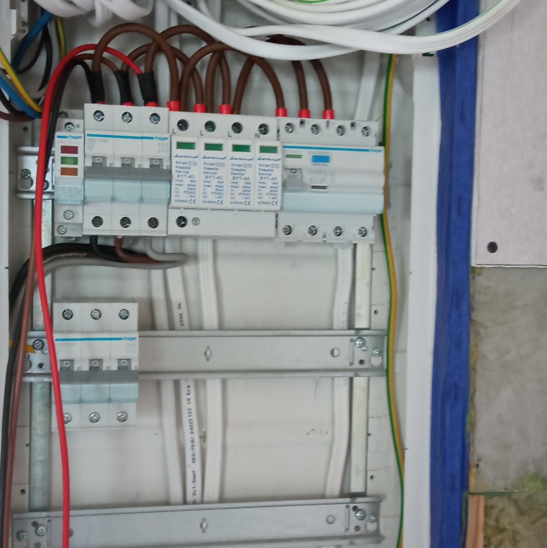 Wnętrze skrzynki elektrycznej z widocznymi zabezpieczeniami przepięciowymi Hager BY7-40, przewodami w brązowej izolacji i elementami montażowymi na szynach.