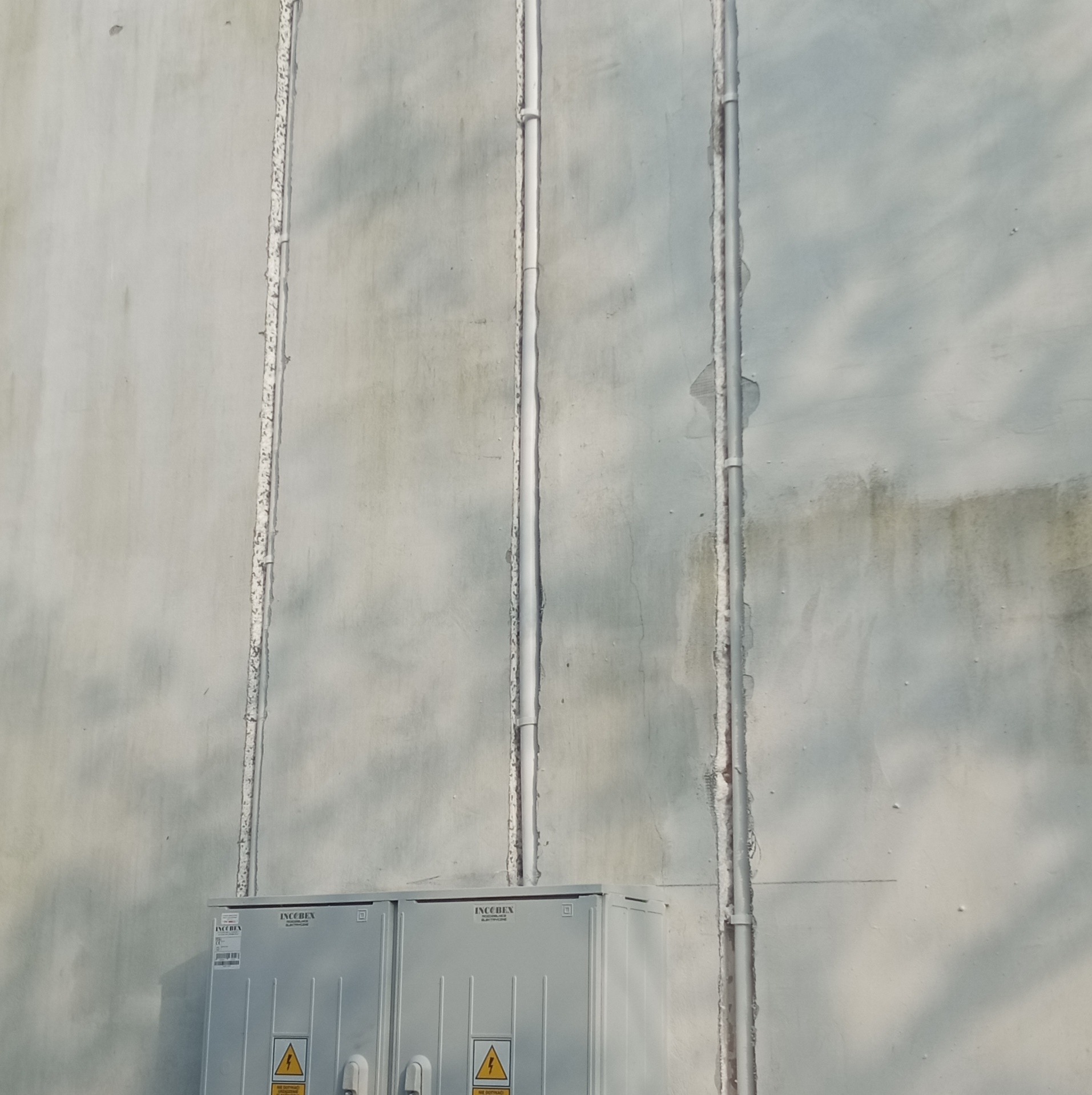 Szara ściana budynku z trzema pionowymi rurami instalacyjnymi i dwoma szarymi skrzynkami elektrycznymi z żółtymi znakami ostrzegawczymi na dole.