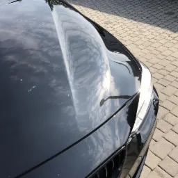 Błyszcząca, czarna maska samochodu odbijająca błękitne niebo z chmurami, widoczne drobne niedoskonałości lakieru i fragment kostki brukowej w tle.