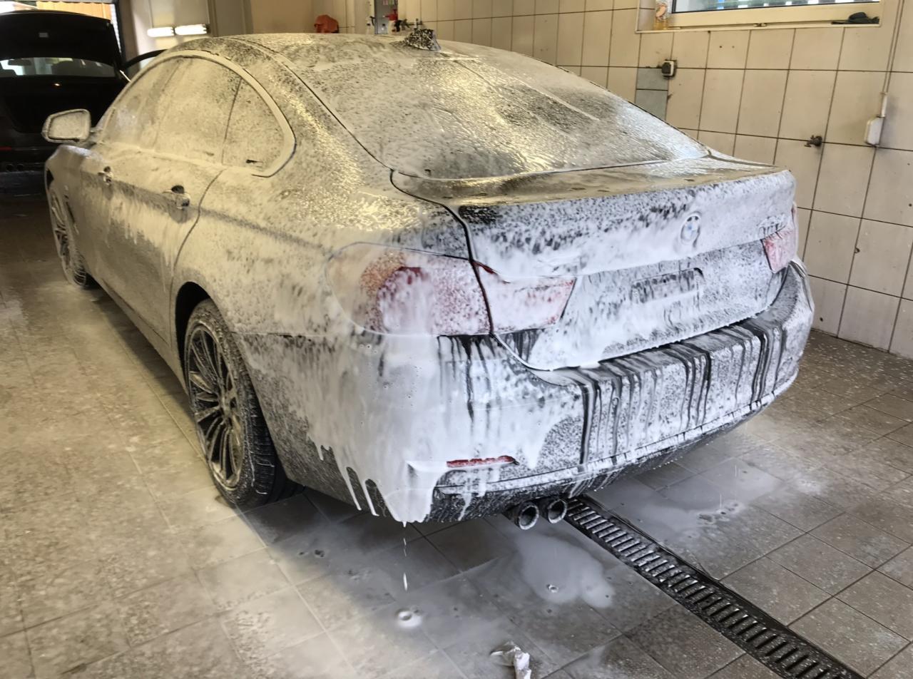 Czarny samochód BMW pokryty pianą podczas mycia wewnątrz myjni samochodowej, widoczne krople spływającej piany i kratka odpływowa.