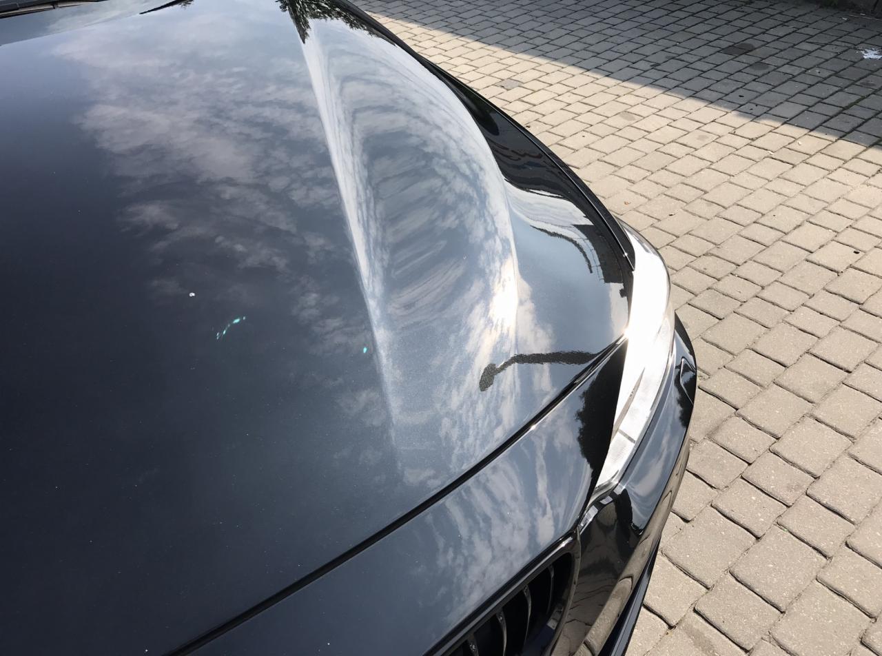 Błyszcząca, czarna maska samochodu odbijająca błękitne niebo z chmurami, widoczne drobne niedoskonałości lakieru i fragment kostki brukowej w tle.