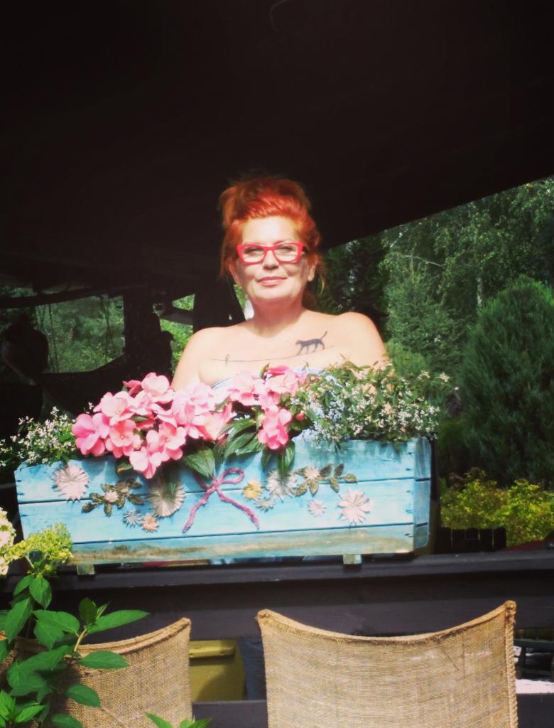 Uśmiechnięta kobieta w czerwonych okularach za skrzynką z kwiatami w Kościelnej Wsi. Widoczny tatuaż kota na ramieniu i bujne, zielone tło.