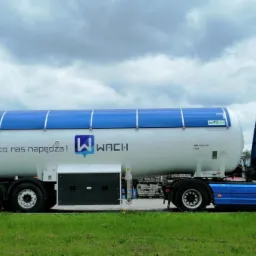 Cysterna paliwowa w barwach firmy WACHI z hasłem 'Paliwa - to co nas napędza!' na tle zielonej trawy i pochmurnego nieba.