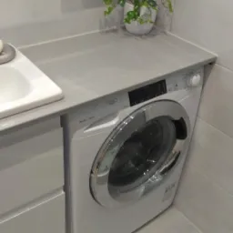 Nowoczesna łazienka z umywalką nablatową, szarą szafką pod umywalką i pralką pod blatem, wykończona jasnymi płytkami.