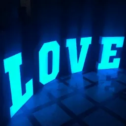 Podświetlane, przestrzenne litery tworzące słowo 'LOVE' w intensywnym, turkusowym kolorze, ustawione na ciemnej, kwadratowej posadzce, odbijające światło.