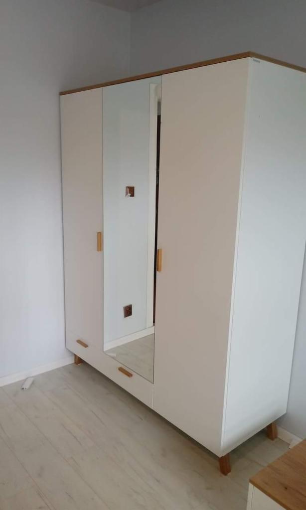 Trzydrzwiowa, biała szafa z lustrem pośrodku i jasnymi, drewnianymi akcentami na uchwytach i nóżkach, ustawiona w rogu jasnego pokoju.