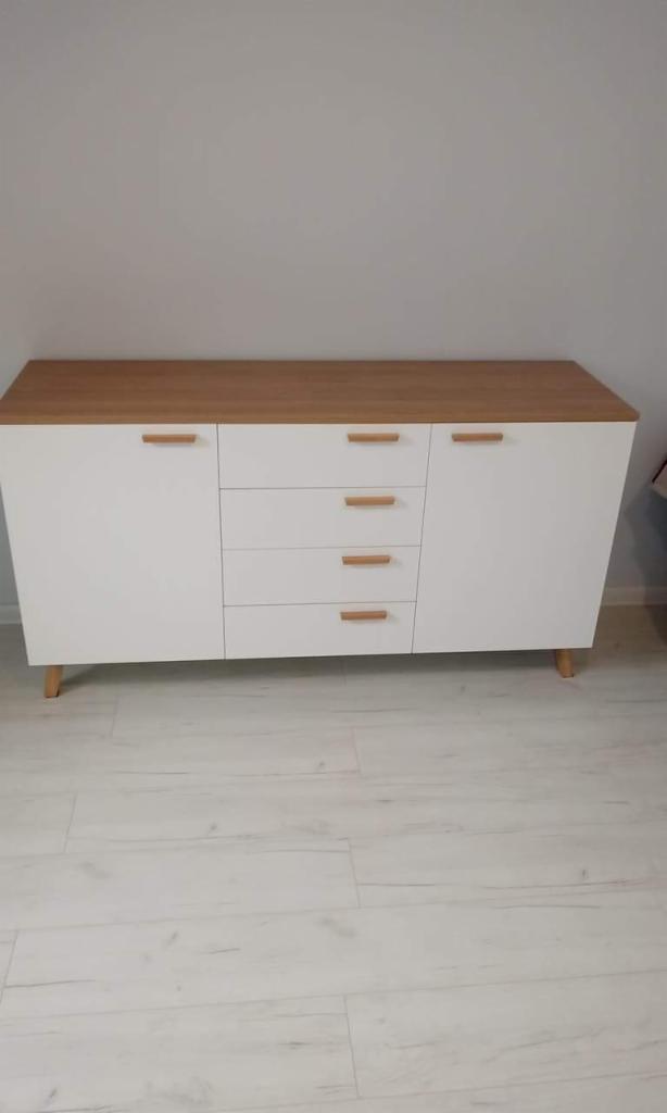 Biała komoda z drewnianym blatem i uchwytami, ustawiona na jasnej podłodze w minimalistycznym wnętrzu.