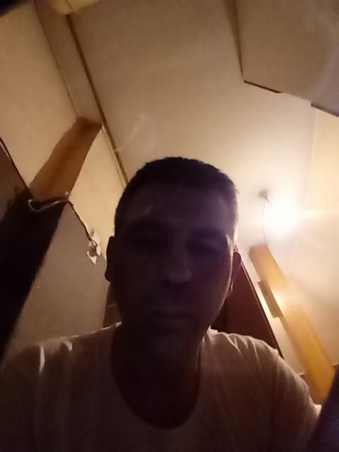 Mężczyzna w słabo oświetlonym pokoju z elementami drewnianej konstrukcji w tle. Ujęcie z dołu, widoczna część sufitu i ściany.