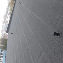 Widok z góry na bitumiczne pokrycie dachu z widocznymi kominkami wentylacyjnymi i budynkami w tle.