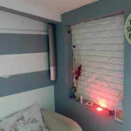 Pokój z dekoracyjną ścianą z białej cegły, podświetloną czerwonym światłem, wazonem z fioletowymi kwiatami, lampą i świecami na parapecie, obok łóżka z poduszką w kwiaty i głośnikiem na ścianie.