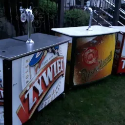 Trzy mobilne bary z kranami do piwa, każdy z logo innej marki: Żywiec, Pilsner Urquell i Tyskie, ustawione na trawniku.