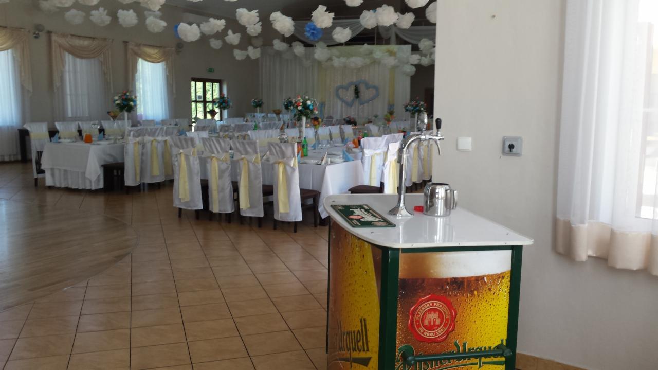 Uroczysta sala weselna z białymi obrusami, krzesłami z żółtymi wstążkami i dekoracją kwiatową, z dystrybutorem piwa na pierwszym planie.