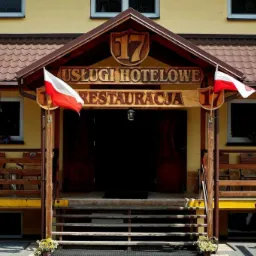 Wejście do budynku z napisem 'Usługi Hotelowe Restauracja' i numerem '17', udekorowane polskimi flagami, schody prowadzą do ciemnych, otwartych drzwi.