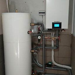 MSS-klima - Biały zbiornik akumulacyjny ciepła z licznymi przyłączami rur i zaworów, obok wiszący na ścianie biały moduł pompy ciepła Gree Versati III z wyświetlaczem, widoczne szare rury instalacyjne...