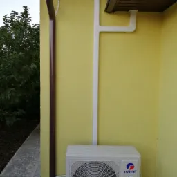 Zewnętrzna jednostka klimatyzatora marki Gree zamontowana na ścianie budynku z żółtą elewacją, obok brązowej rynny i białej instalacji odprowadzającej skropliny.