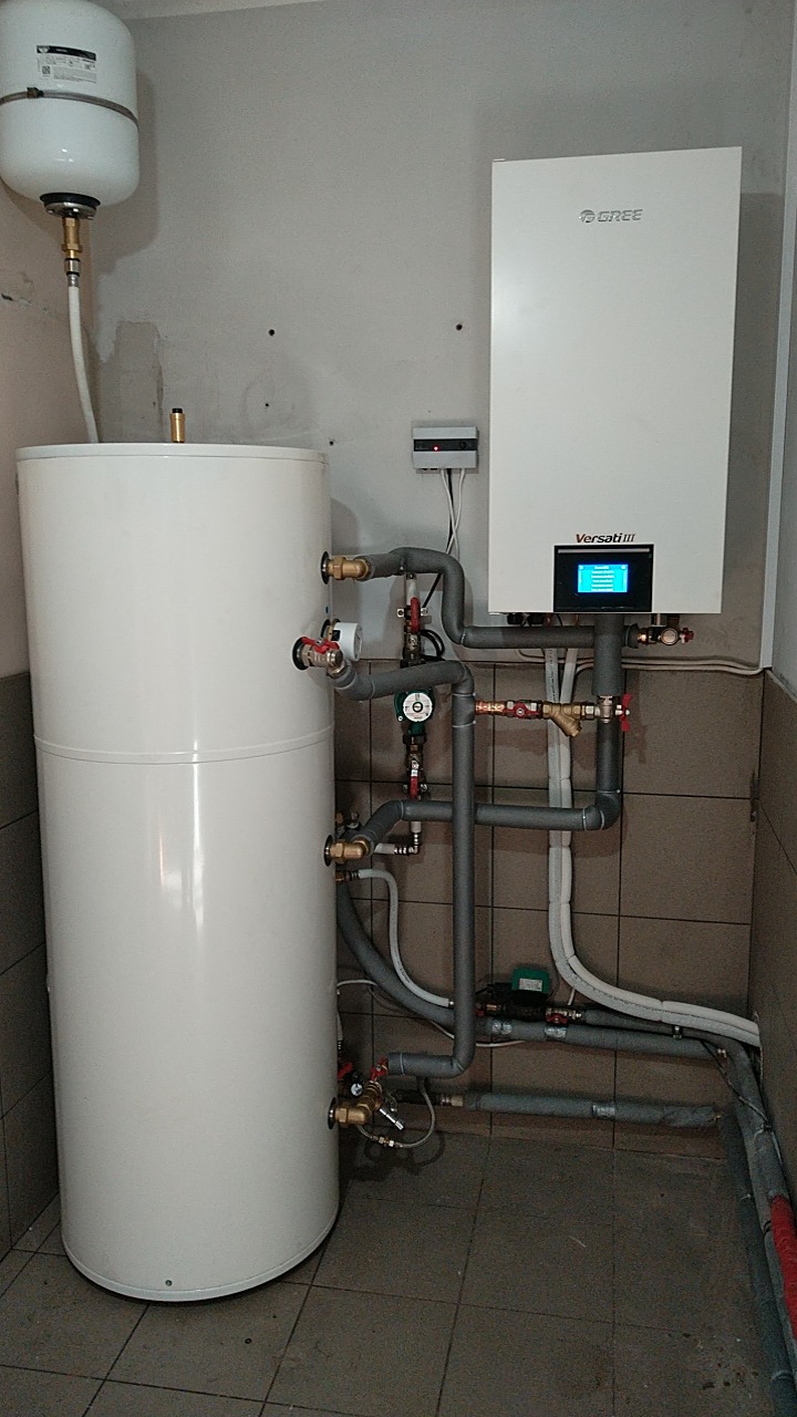 Biały zbiornik akumulacyjny ciepła z licznymi przyłączami rur i zaworów, obok wiszący na ścianie biały moduł pompy ciepła Gree Versati III z wyświetlaczem, widoczne szare rury instalacyjne...