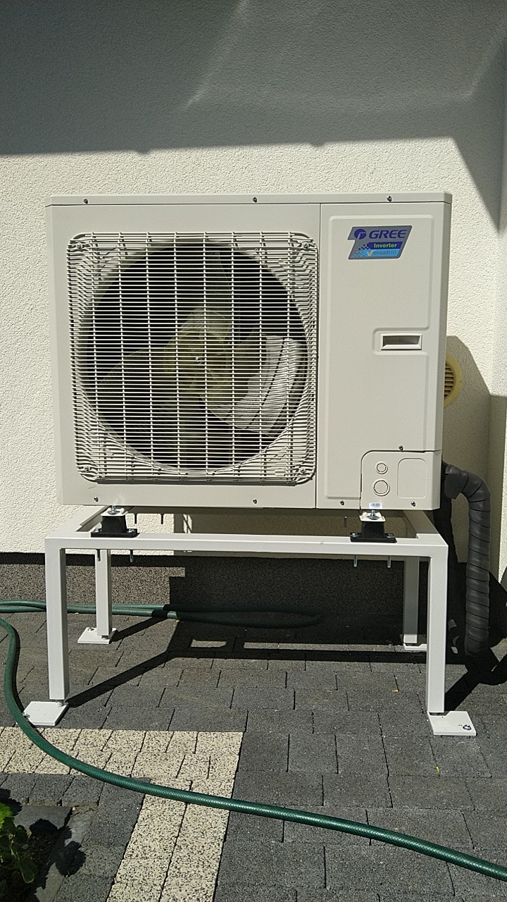 Zewnętrzna jednostka klimatyzacji Gree Inverter Versati III zamontowana na białym, metalowym stelażu na tle jasnej ściany budynku i szarej kostki brukowej. Widoczny fragment zielonego węża ogrodowego.