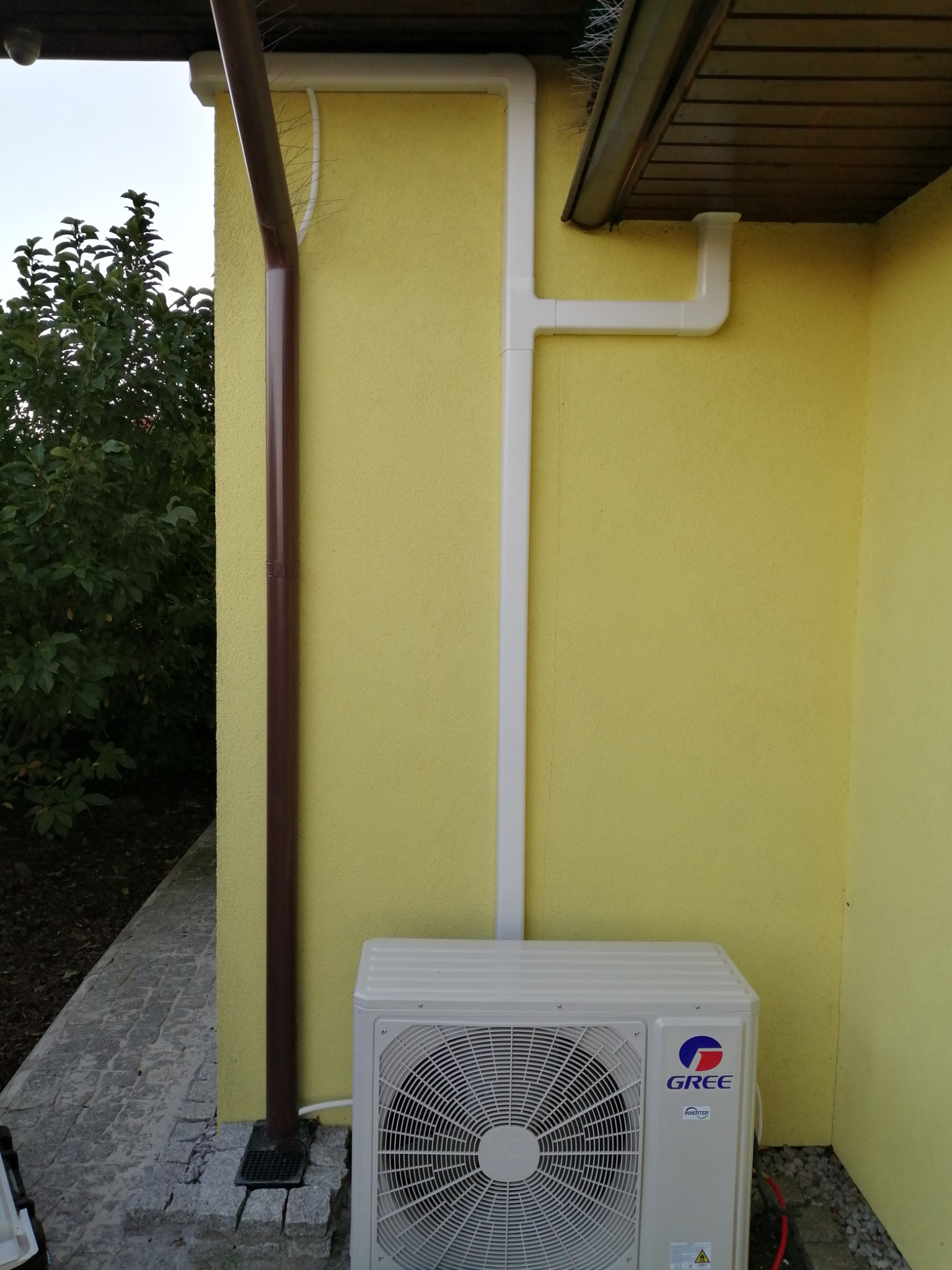 Zewnętrzna jednostka klimatyzatora marki Gree zamontowana na ścianie budynku z żółtą elewacją, obok brązowej rynny i białej instalacji odprowadzającej skropliny.