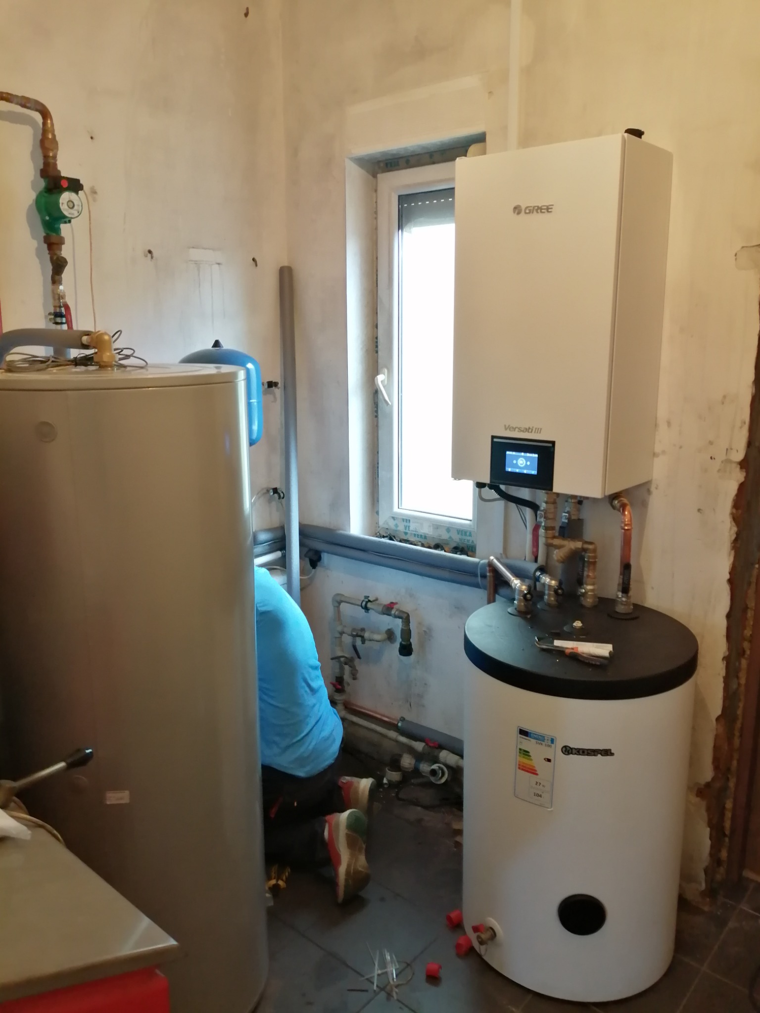 Montaż pompy ciepła Gree Versati III w surowym pomieszczeniu, widoczne rury, zbiornik Kostrzewa i instalator w niebieskiej koszulce.