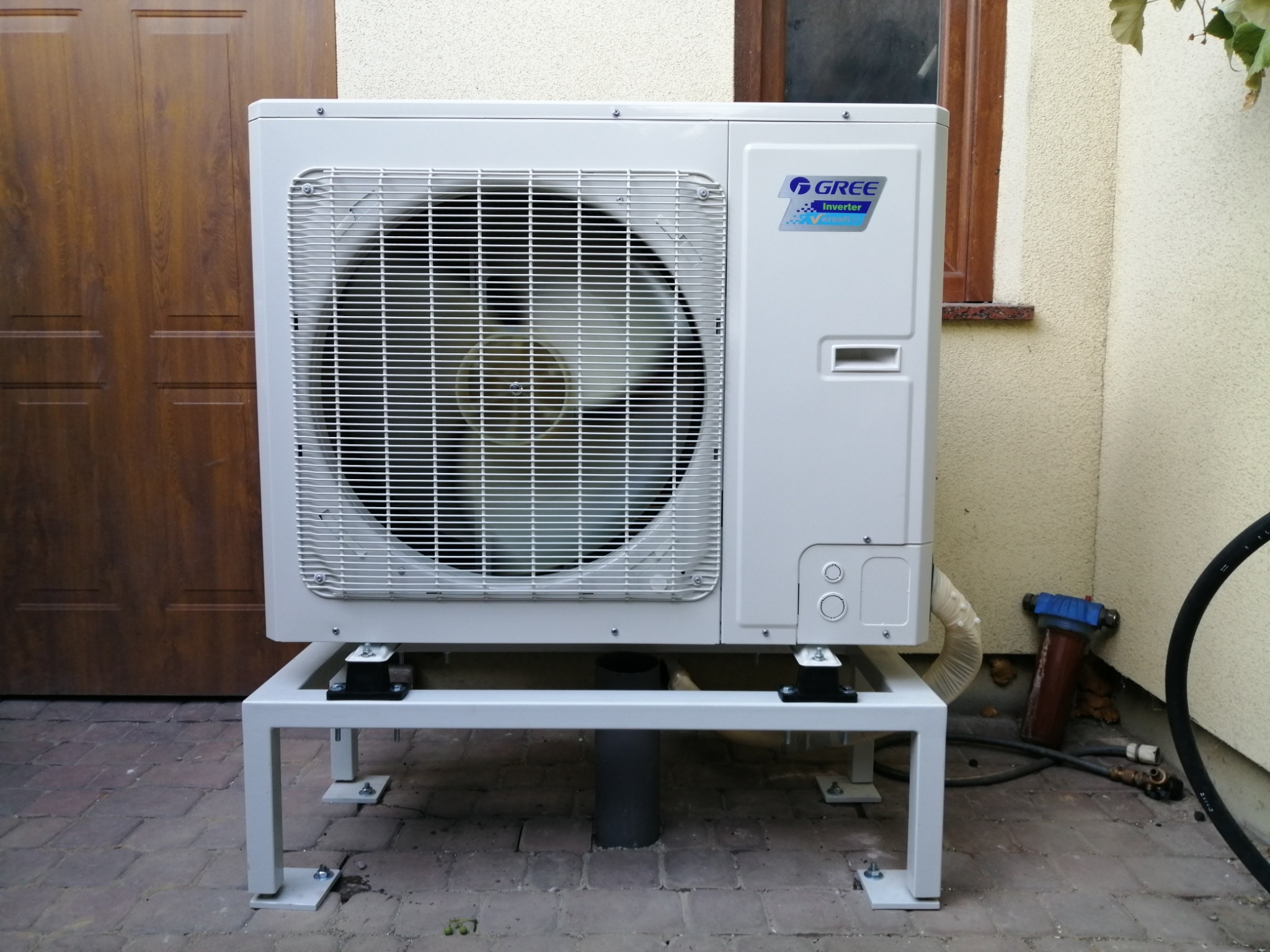 Biała zewnętrzna jednostka pompy ciepła Gree zamontowana na metalowej podstawie, zainstalowana przy ścianie budynku w Oławie, widoczny filtr wody i przewody.