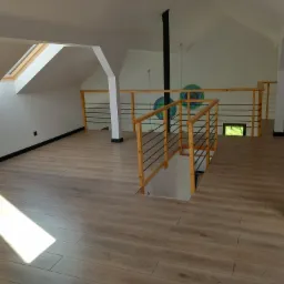 Jasne, przestronne poddasze z drewnianą podłogą, widoczne schody z drewnianą balustradą z czarnymi prętami, okno dachowe i dekoracyjne, okrągłe obrazy na ścianie.