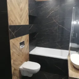 Nowoczesna łazienka z czarnymi płytkami imitującymi marmur, drewnopodobnymi płytkami ułożonymi w jodełkę, podwieszaną toaletą i białą wanną z parawanem.