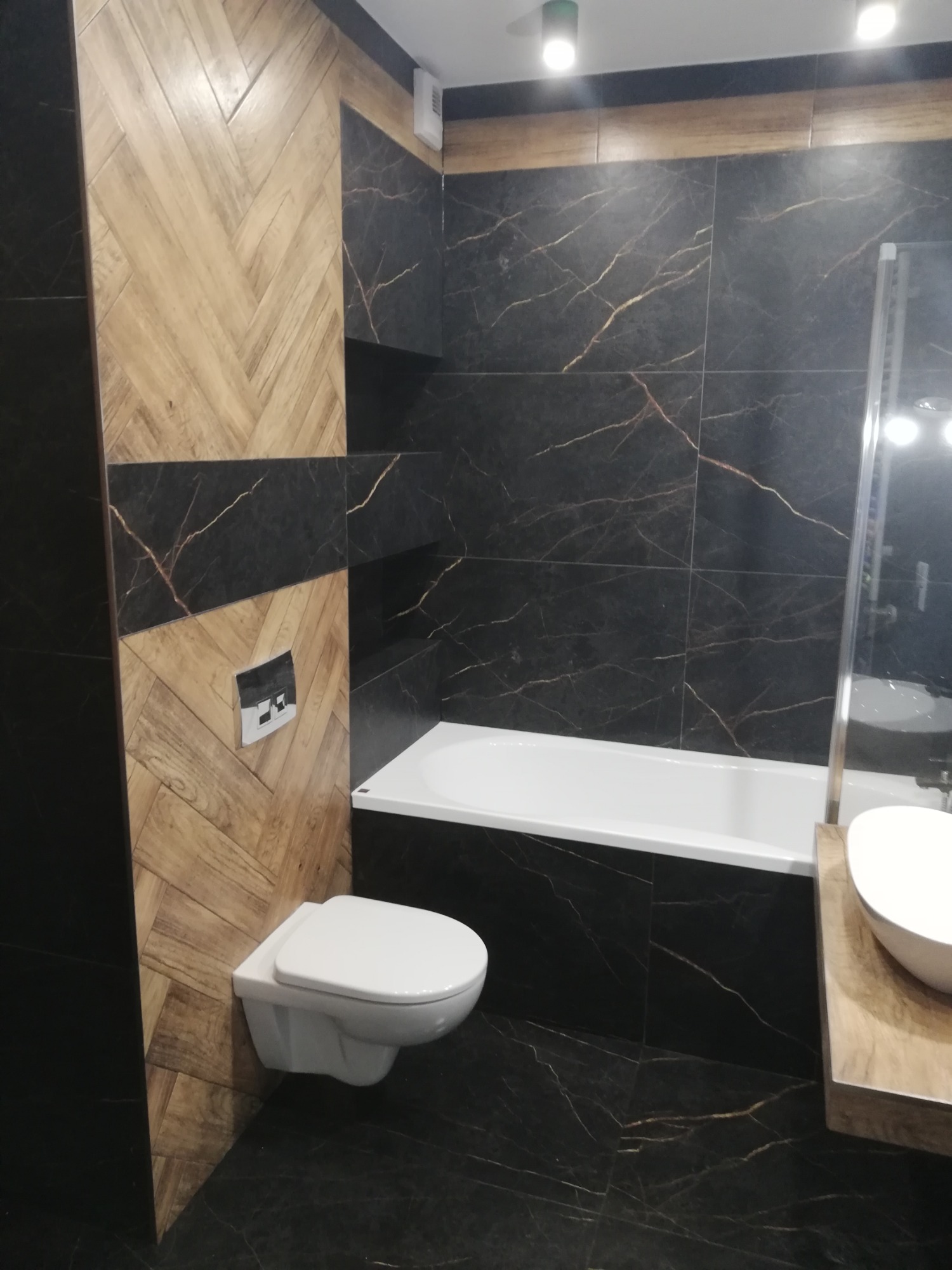 Nowoczesna łazienka z czarnymi płytkami imitującymi marmur, drewnopodobnymi płytkami ułożonymi w jodełkę, podwieszaną toaletą i białą wanną z parawanem.