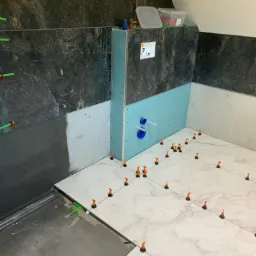 Montaż płytek imitujących marmur w łazience z użyciem systemu poziomowania; widoczne fragmenty ściany z czarnego gresu i płyty karton gips.