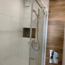 Kabina prysznicowa z chromowanymi okuciami i deszczownicą, białe płytki na ścianach, akcent z płytek imitujących drewno oraz ukryty zawór rewizyjny w ramce.