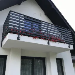 Czarna, pozioma balustrada balkonowa z metalowych profili, ozdobiona czerwonymi kwiatami, na tle białej ściany domu z ciemnym dachem.