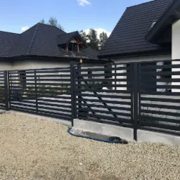 Nowoczesne, poziome przęsła w kolorze grafitowym z furtką, tworzące ogrodzenie posesji z dwoma domami w tle, widoczne przyłącze elektryczne i rozsypany żwir.
