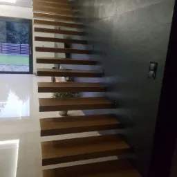 Nowoczesne schody drewniane wspornikowe, zamocowane do ciemnej ściany, z widocznym oknem i fragmentem ogrodzenia w tle.