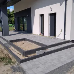 Nowoczesny dom z szarą kostką brukową na podjeździe i schodach, z przygotowanym miejscem na rabatę kwiatową wypełnioną piaskiem, widoczne narzędzia i przewód doprowadzający wodę.