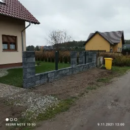 Wznoszony z szarych bloczków betonowych niski murek ogrodzeniowy z grafitowymi słupkami, widok z boku na posesję z dwoma domami i żółtym pojemnikiem na śmieci w tle, pochmurny dzień.