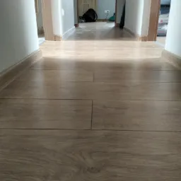 Ułożone panele podłogowe w korytarzu, widok wzdłuż pomieszczenia, z widocznymi listwami przypodłogowymi i otwartymi drzwiami w tle.