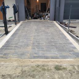 Przem-Bud - Układanie szarej kostki brukowej na podjeździe do garażu, widoczne narzędzia i materiały budowlane w tle, pracownik z miotłą w lewym górnym rogu.