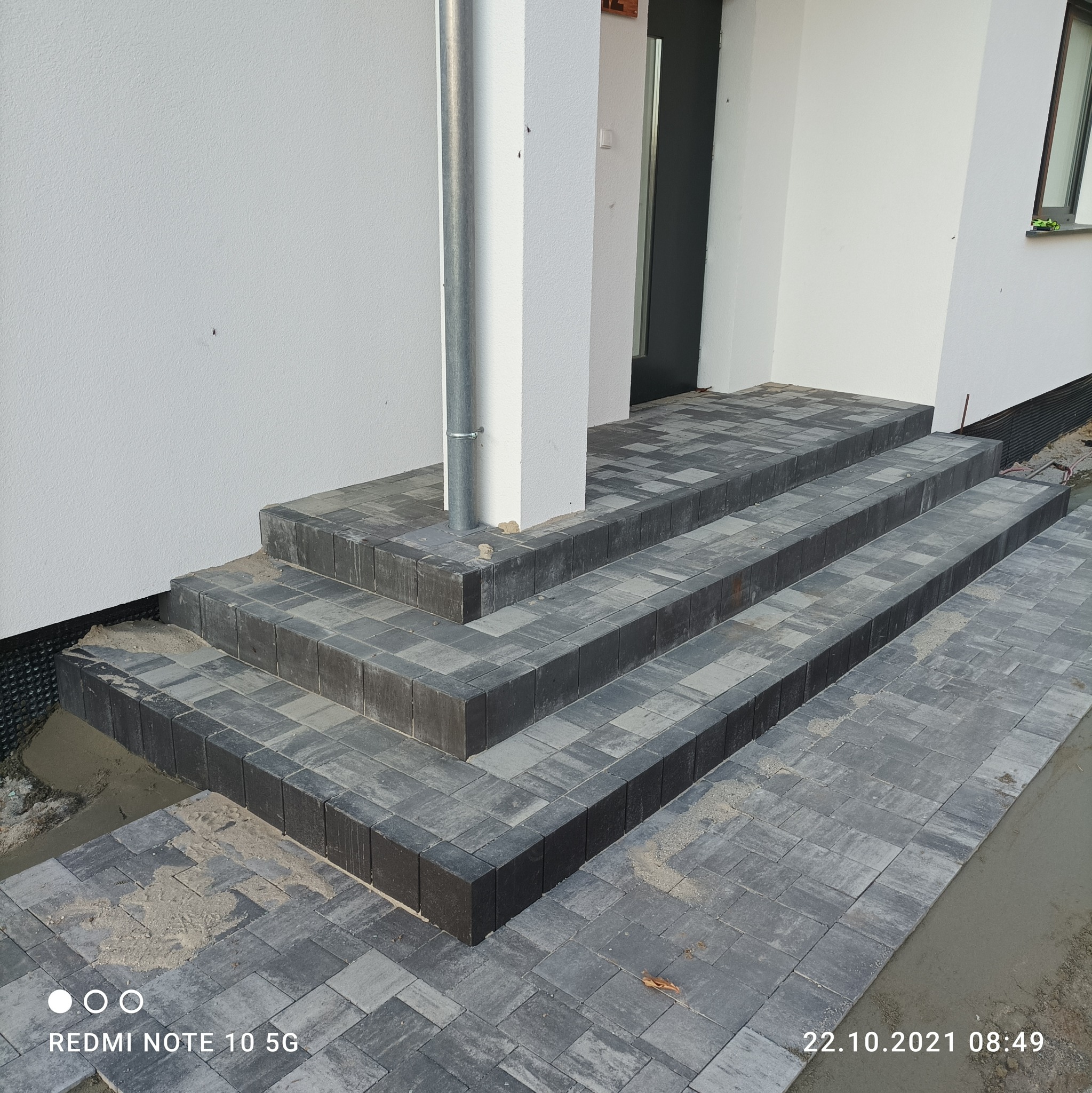 Nowe schody wejściowe wyłożone szarą kostką brukową prowadzące do domu z białą elewacją, widoczna rynna i fragment okna.
