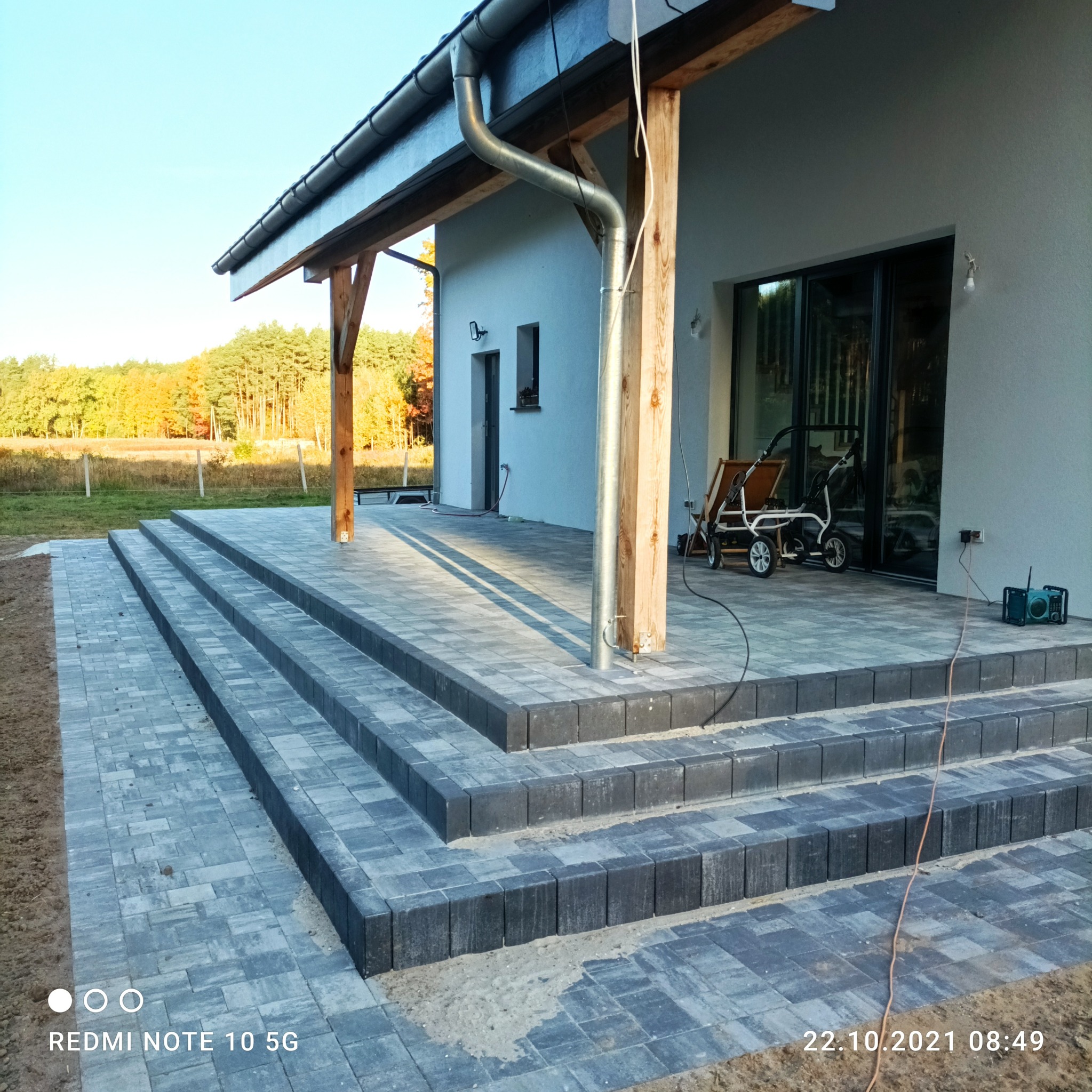 Nowoczesny taras z szarej kostki brukowej, schody prowadzące do domu z drewnianą konstrukcją zadaszenia, widok na jesienny las w tle.