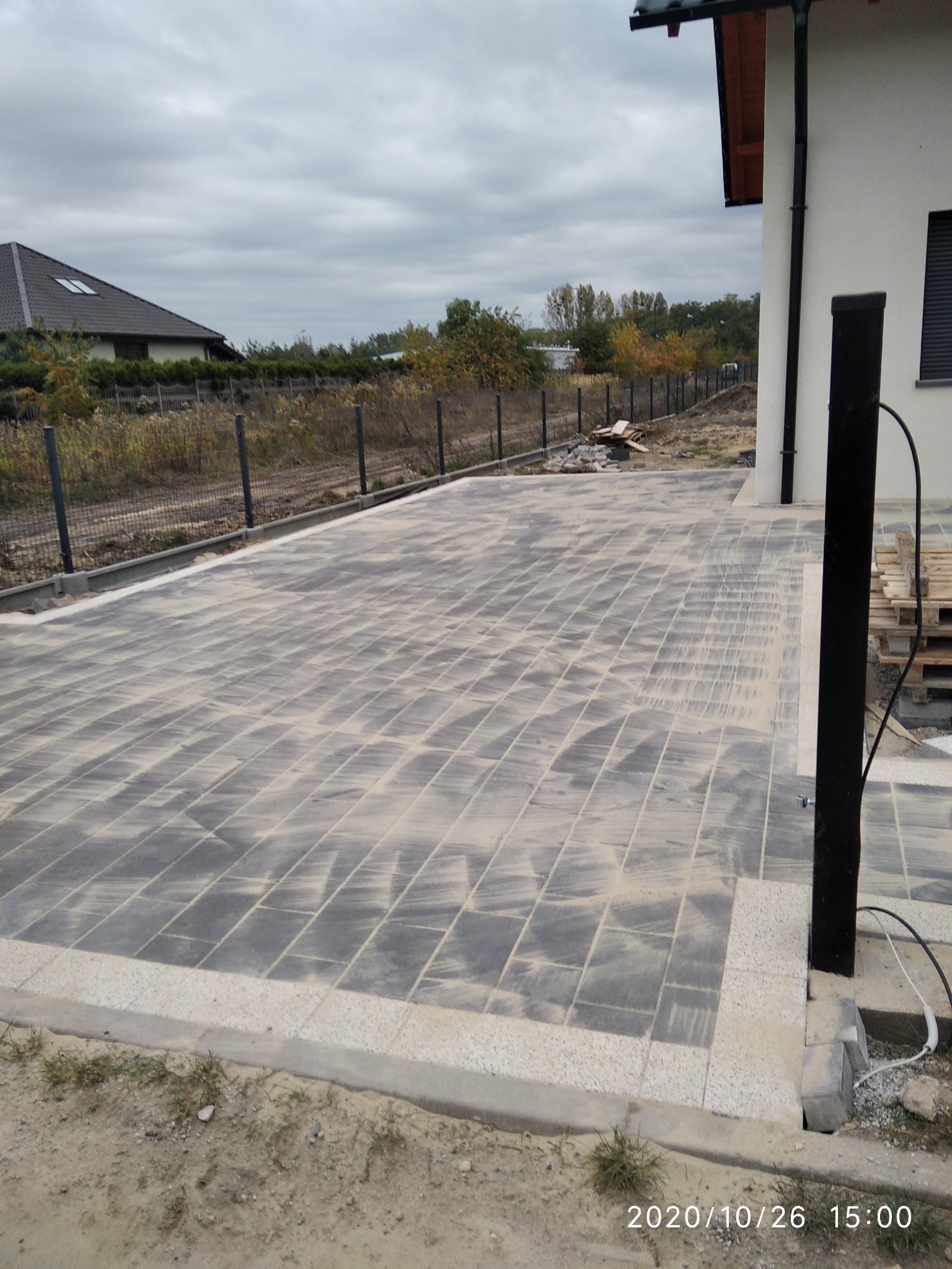 Ułożona kostka brukowa na podwórku, widoczne słupki ogrodzeniowe w tle, palety drewniane przy ścianie budynku.