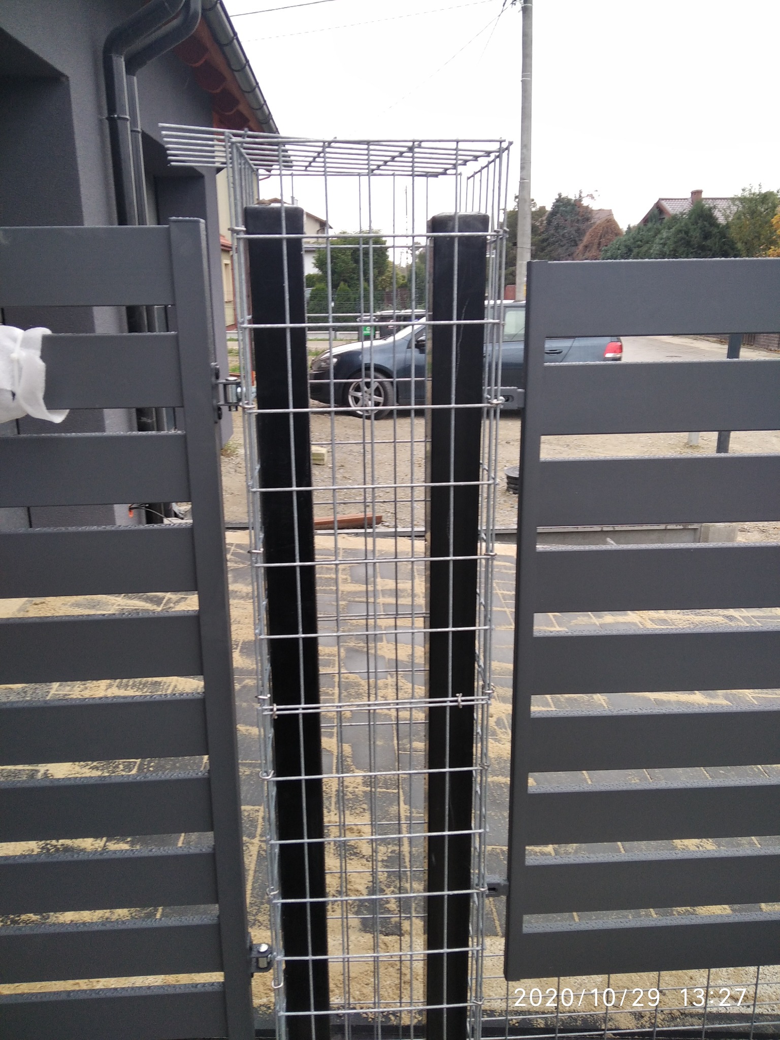 Nowoczesna, grafitowa brama wjazdowa z metalowymi słupkami wypełnionymi siatką gabionową, widok z zewnątrz posesji.