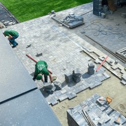 Your Smart Garden Sp. z o.o. - Układanie szarej kostki brukowej na posesji, widok z góry, pracownicy w zielonych koszulkach, palety z kostką brukową, trawnik w tle.
