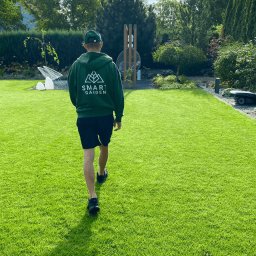 Your Smart Garden Sp. z o.o. - Mężczyzna w zielonej bluzie z logo firmy spaceruje po idealnie przystrzyżonym, zielonym trawniku w ogrodzie. W tle widoczne drzewa i elementy wyposażenia ogrodu, w tym robot koszący.