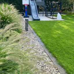 Nowoczesny ogród z domkiem na drzewie, zjeżdżalnią i boiskiem do koszykówki, obrzeża z kamieni i ozdobne trawy, zielony trawnik.