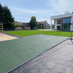 Your Smart Garden Sp. z o.o. - Przygotowanie podłoża pod plac zabaw z gumowych płyt, widoczna czarna folia, w tle trawnik i nowoczesny dom.