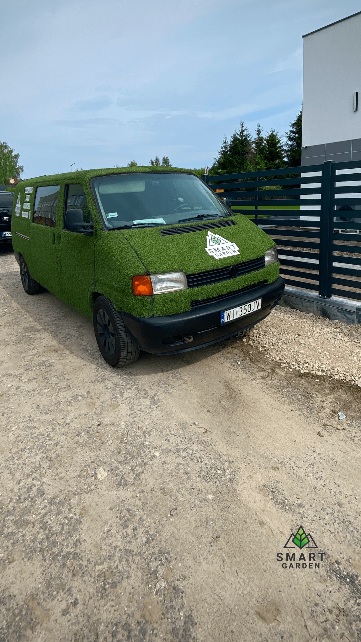 Samochód dostawczy oklejony sztuczną trawą z logo Smart Garden zaparkowany na żwirowym podłożu, w tle nowoczesny dom z ciemnym ogrodzeniem.