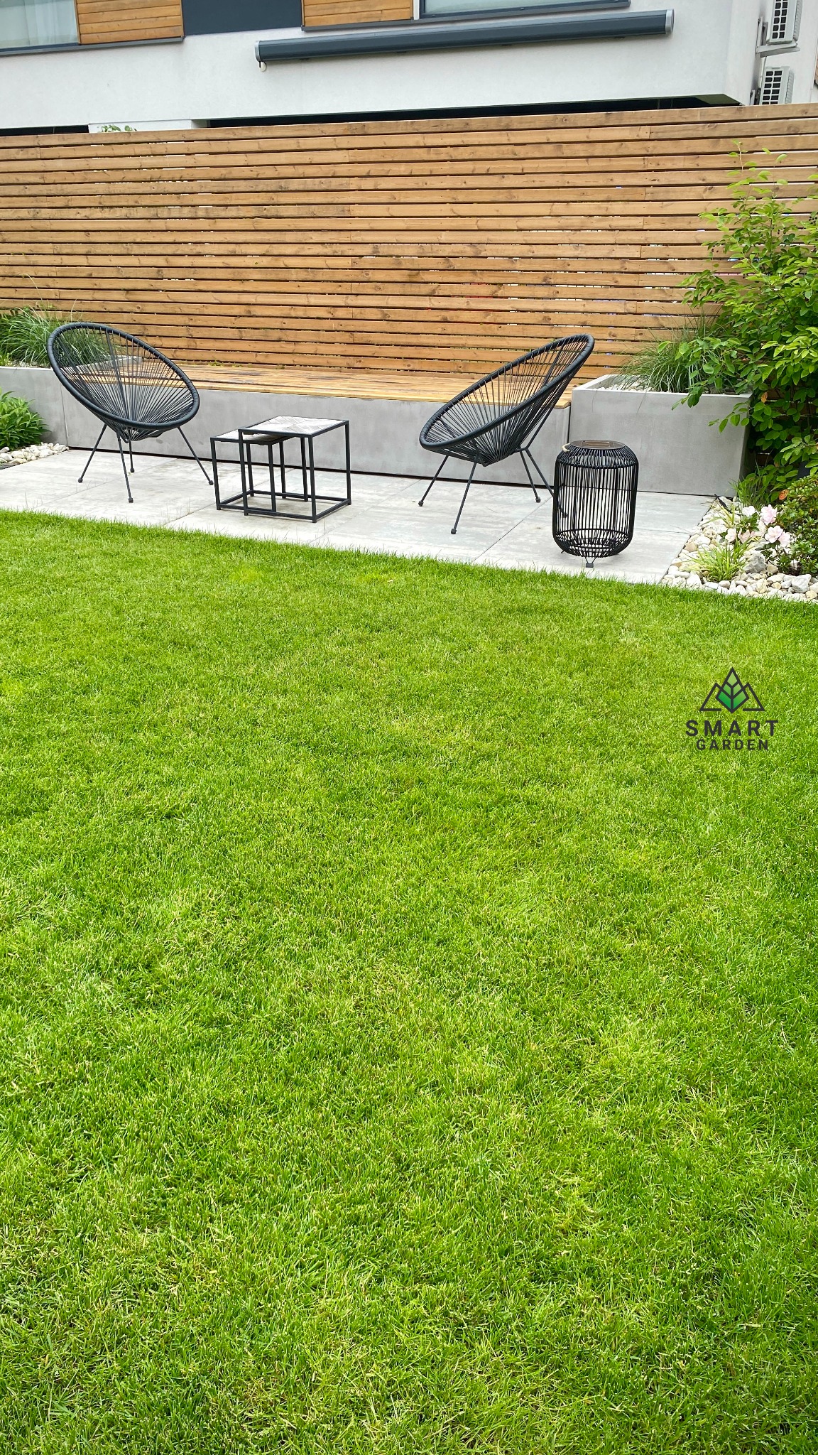 Nowoczesny ogród z równo przystrzyżonym trawnikiem, dwoma czarnymi fotelami z plecionki, stolikiem kawowym i drewnianym panelem ogrodzeniowym w tle.