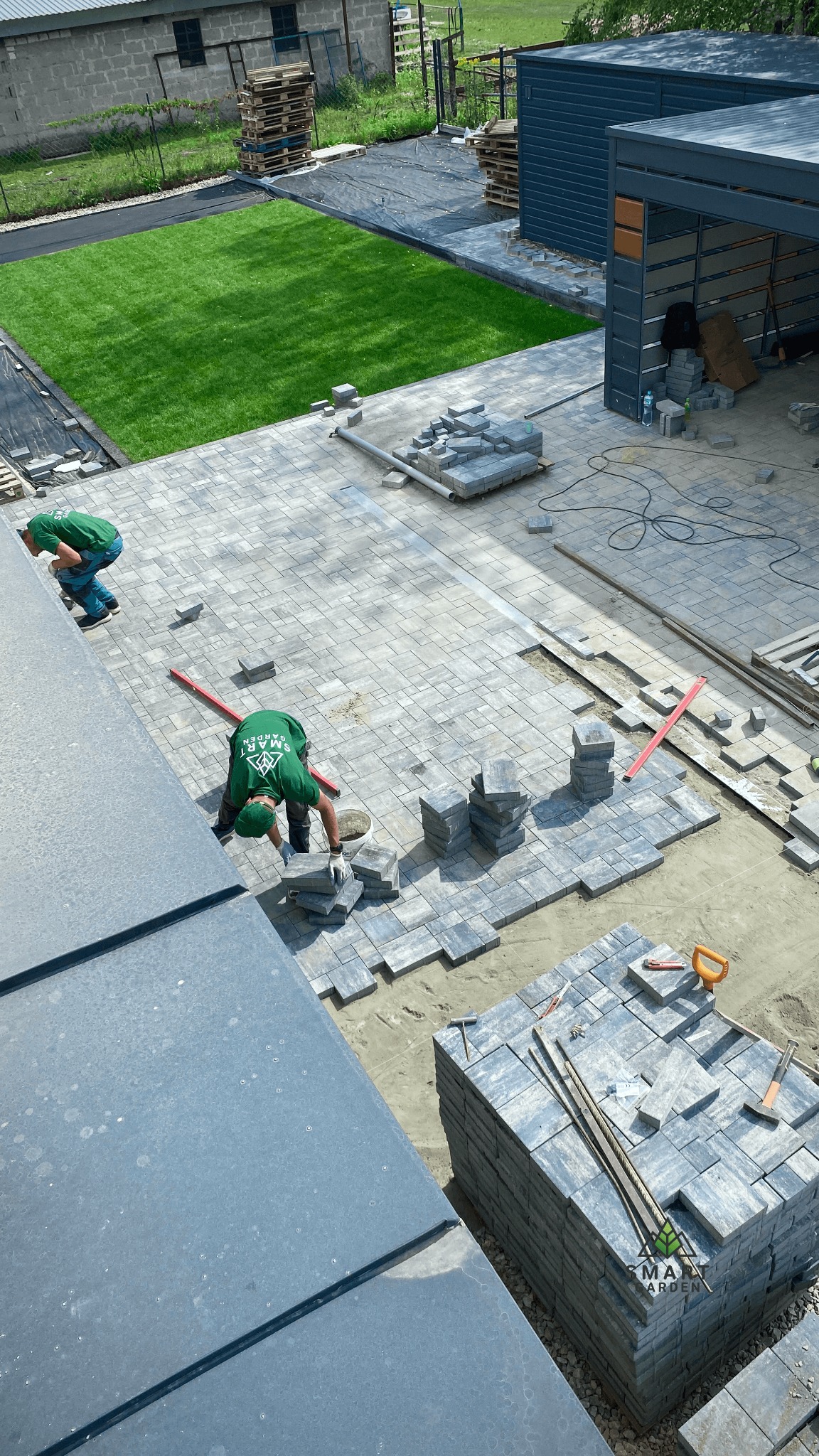 Układanie szarej kostki brukowej na posesji, widok z góry, pracownicy w zielonych koszulkach, palety z kostką brukową, trawnik w tle.